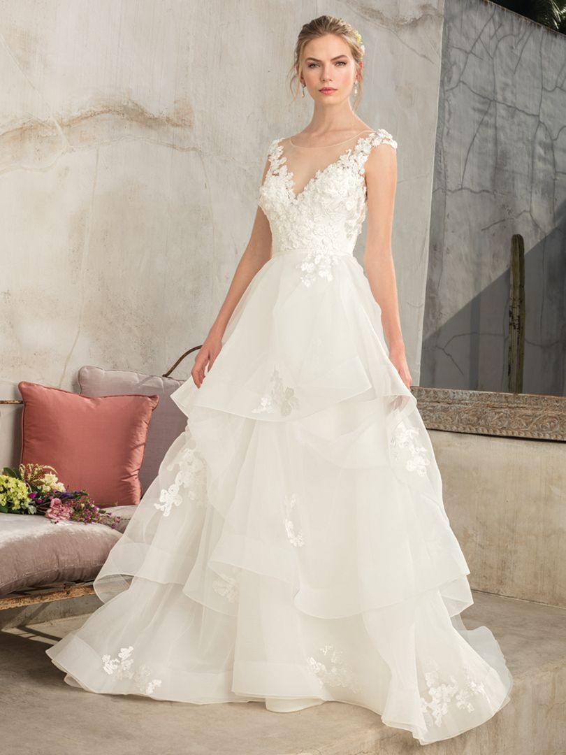 Woman in a white wedding dress with a tiered skirt, standing outside with a light background.