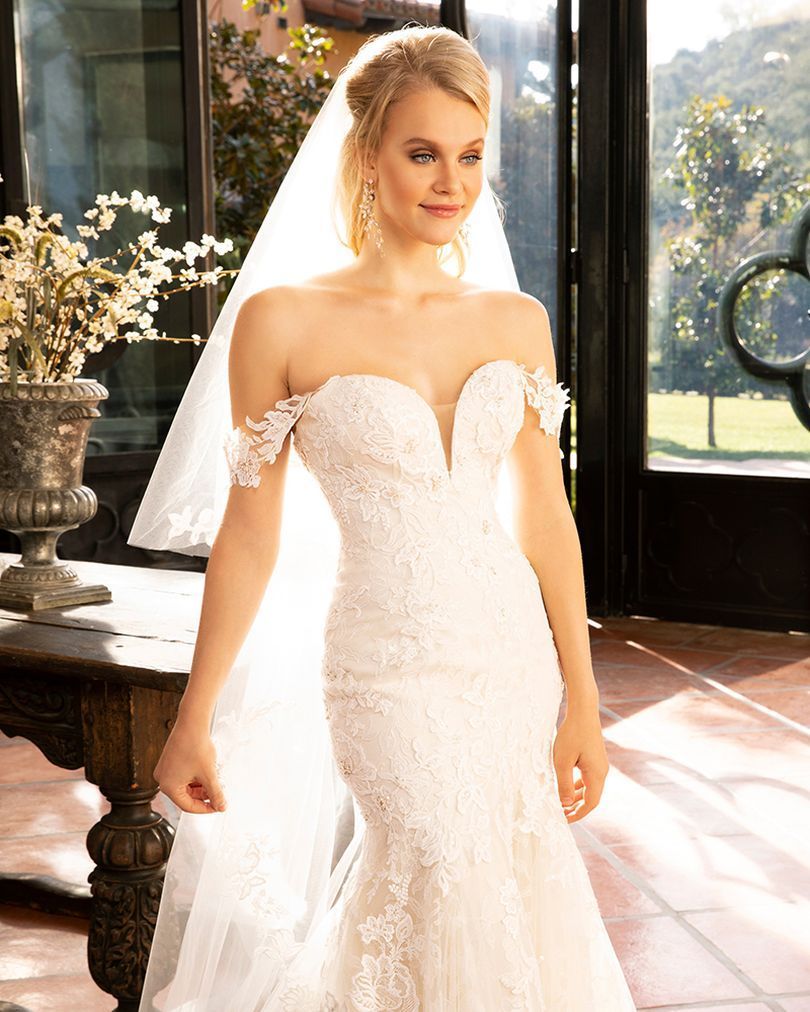 Woman in a white wedding dress with a long train and veil. Standing in front of a window.