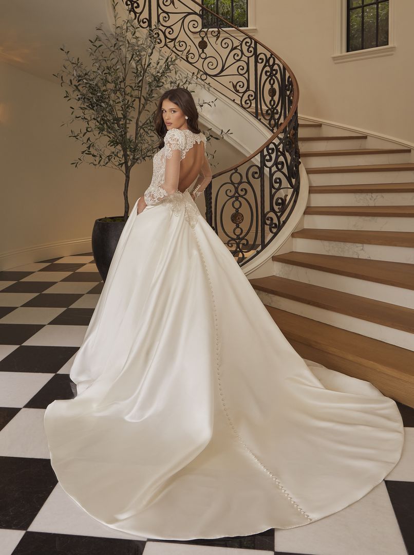 Woman in a wedding gown poses on a checkered floor by a grand staircase with a trailing skirt.