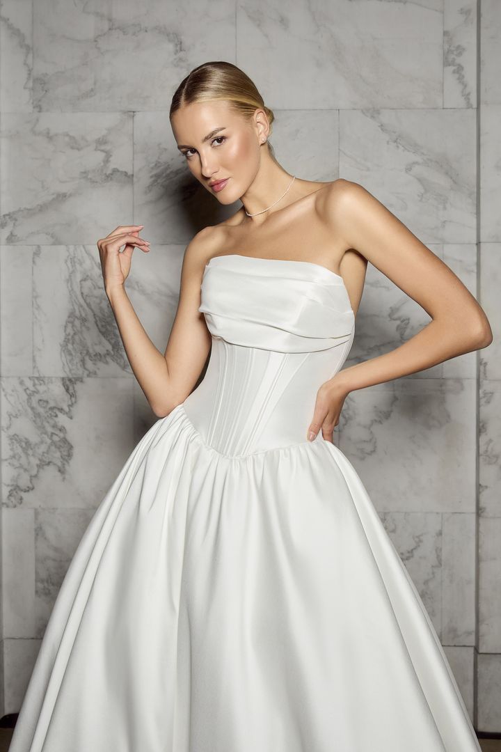 Woman in a white satin wedding gown, posing against a marble wall.