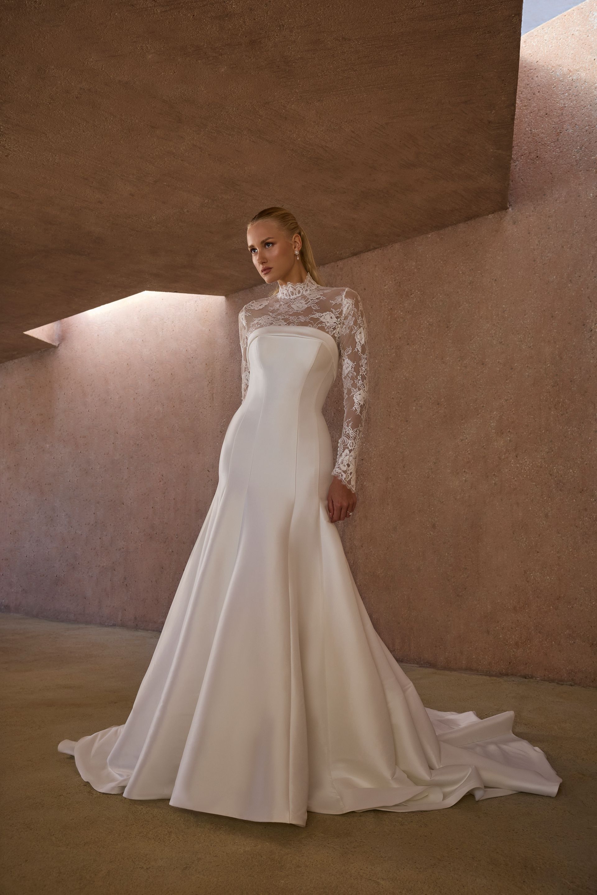 Woman in a white wedding gown with lace sleeves and a train, standing in a textured brown space.