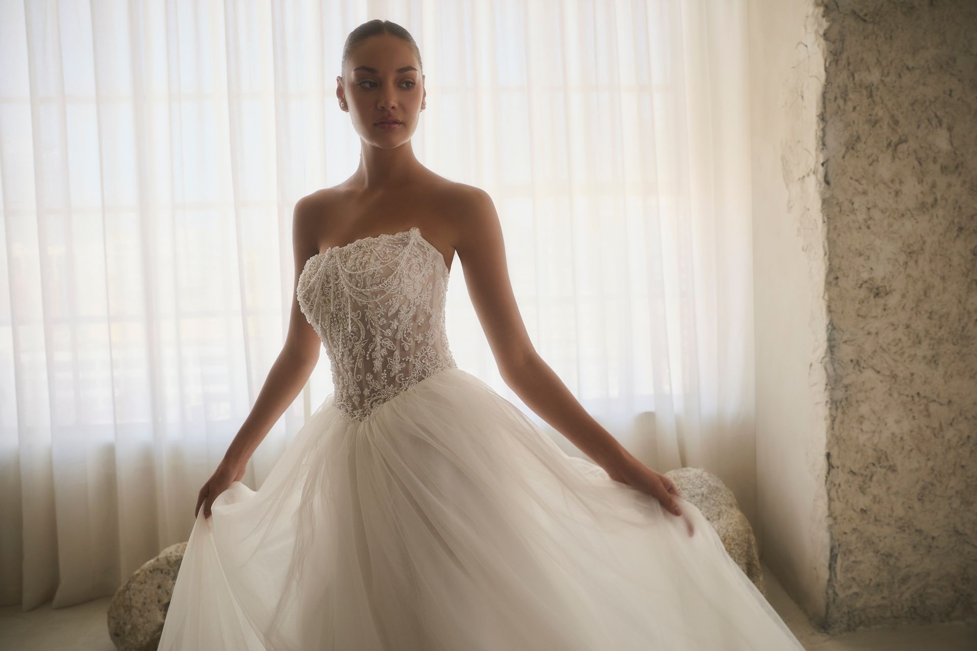 Woman in a strapless white wedding dress stands near a window, holding the skirt out.