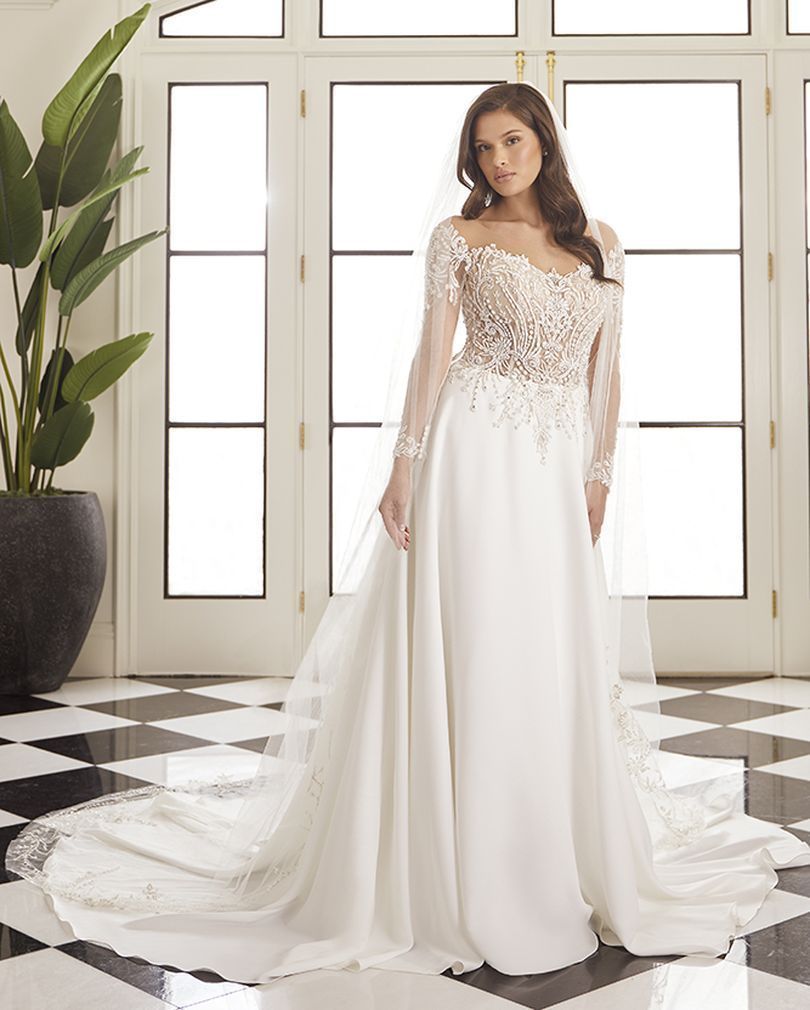 Woman in a white wedding dress with lace bodice, long sleeves, and a flowing skirt, standing in front of a window.