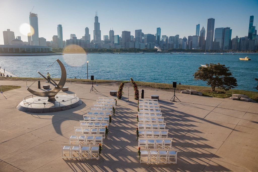 Wedding ceremony setup on a waterfront with Chicago skyline in the background. Rows of white chairs face an arch.