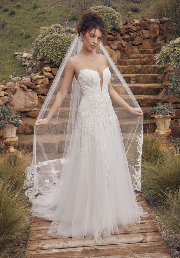 Woman in strapless wedding dress with veil, standing on wooden path, outdoor setting.