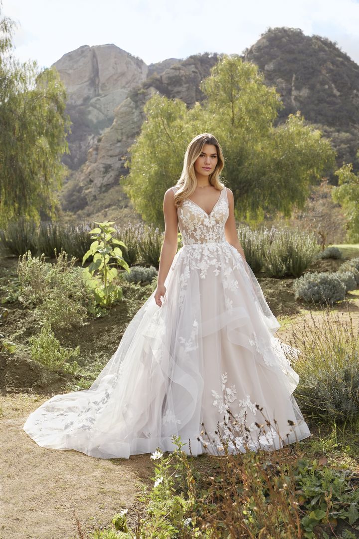 Woman in white wedding dress with floral bodice and flowing skirt, outdoors in a garden setting.