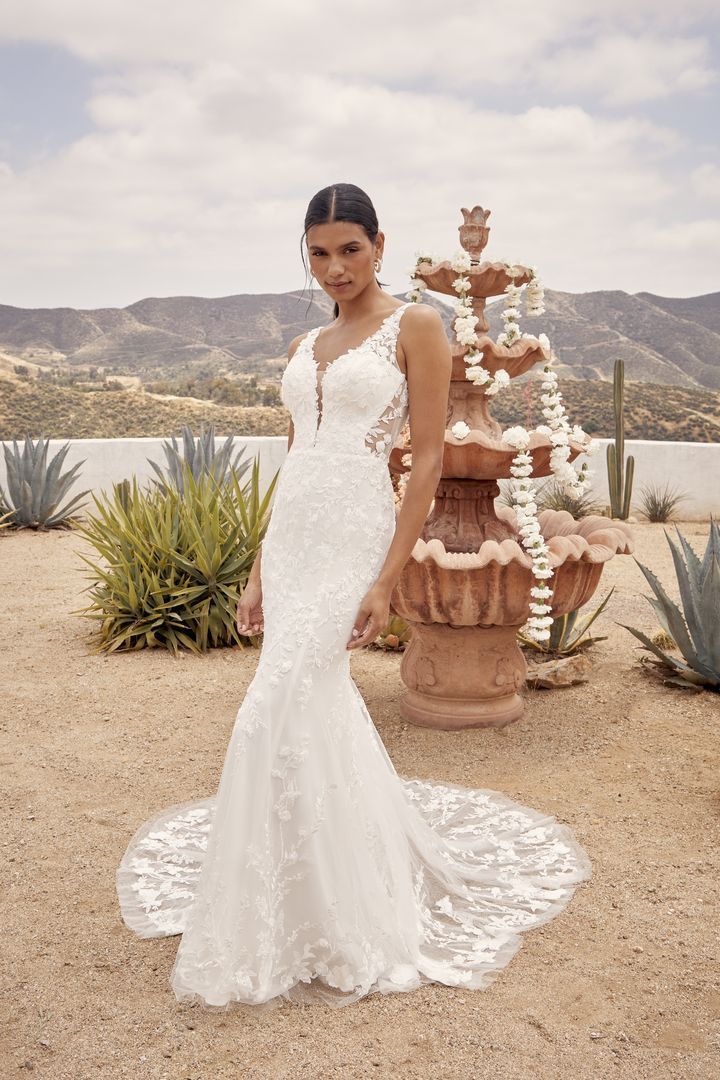 Woman in a white wedding dress stands near a fountain in an outdoor desert setting.