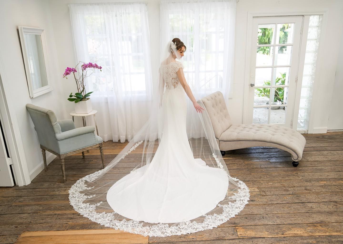 Bride in a lace-trimmed veil and wedding dress in a bright room with a chaise lounge and open door.
