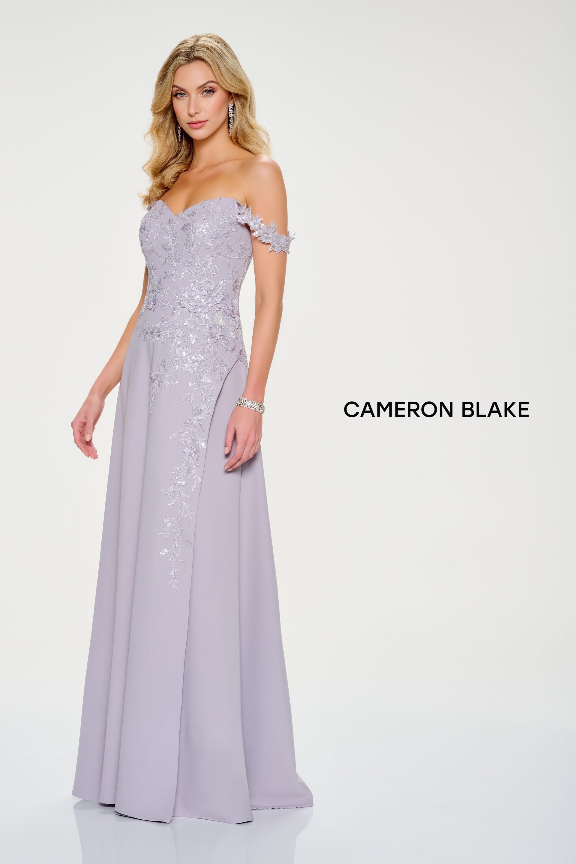 Woman in an off-the-shoulder lavender gown with beaded bodice and flowing skirt, posing in a studio.