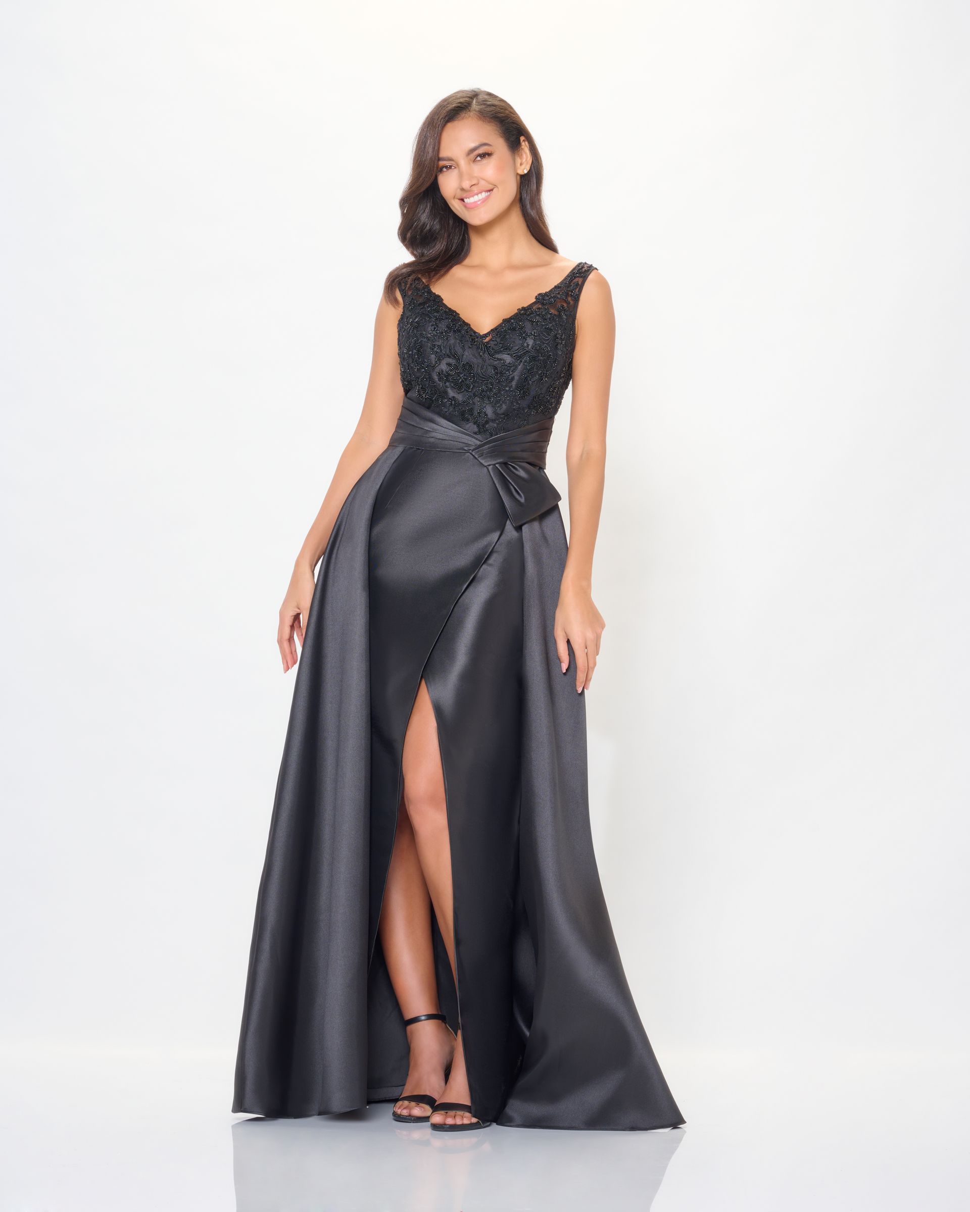 Woman in a formal black dress with beaded bodice, slit skirt, and high heels, smiling, against white background.