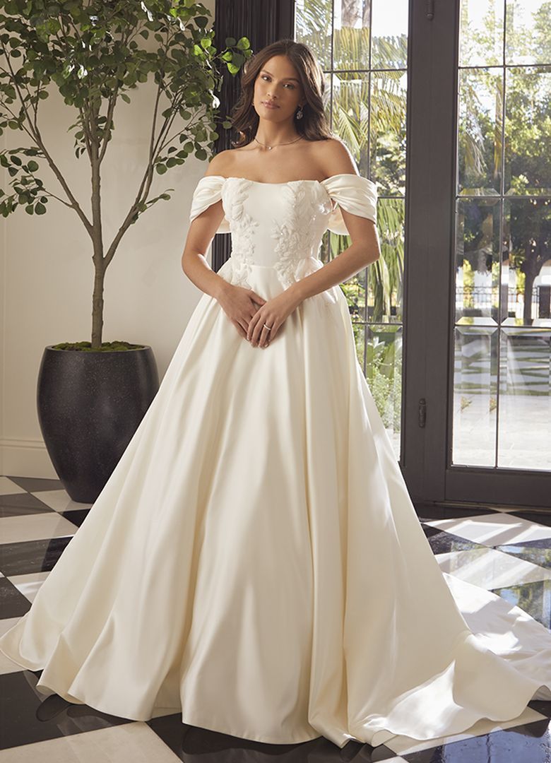 Woman in off-shoulder satin wedding dress, standing indoors near a large tree and window.