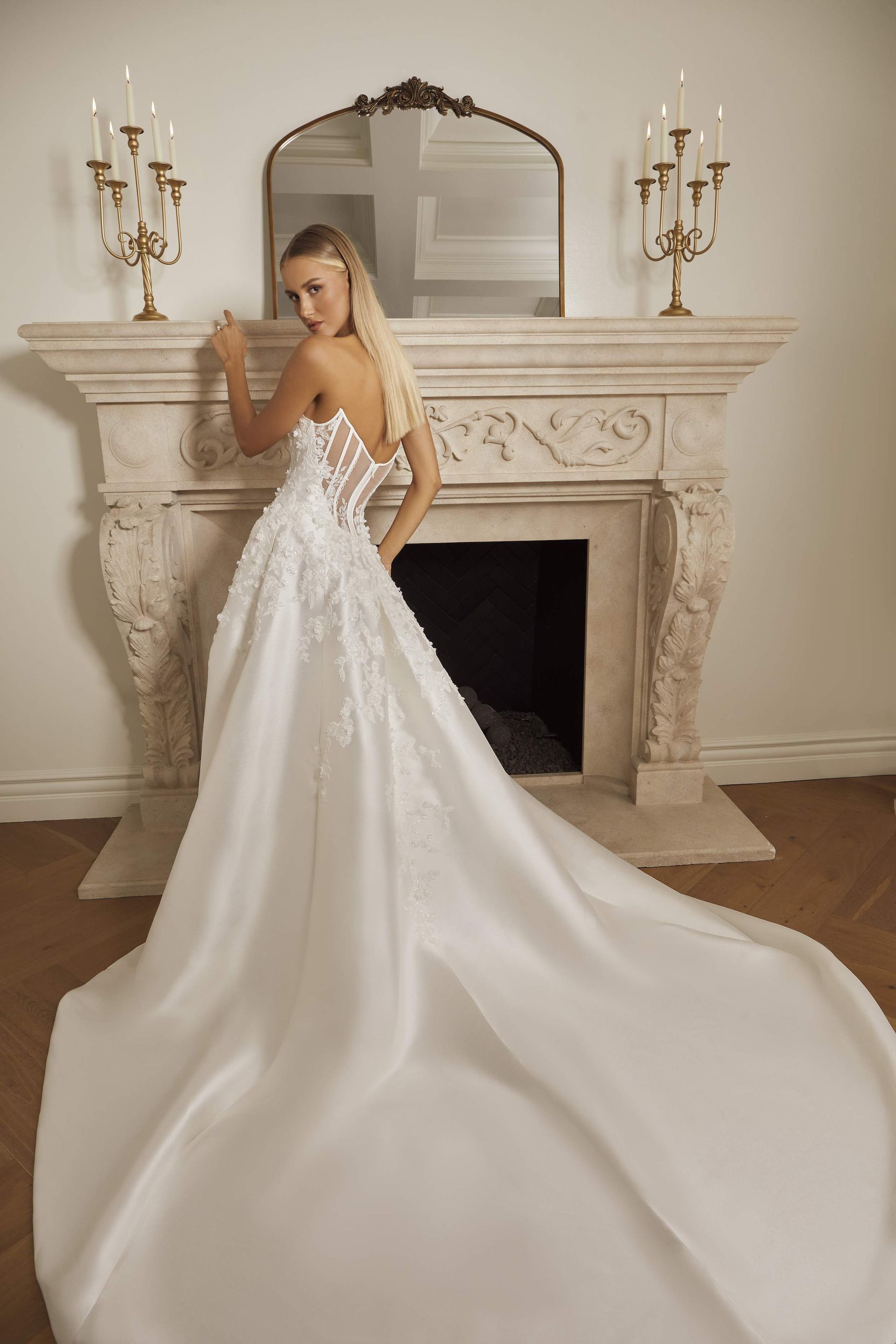 Woman in a white wedding gown leans against a fireplace, the dress train sweeping the floor.
