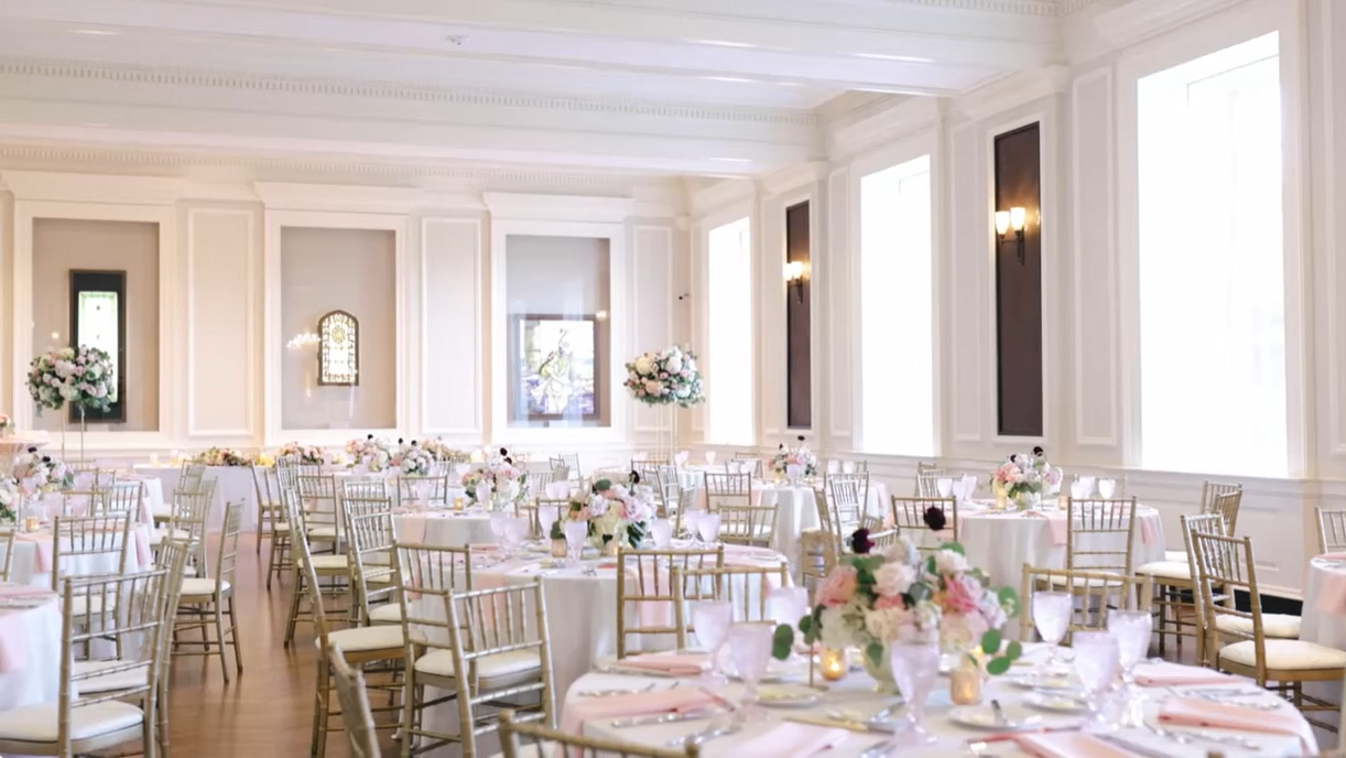 Elegant ballroom set for a wedding, with tables, floral centerpieces, and gold chairs.