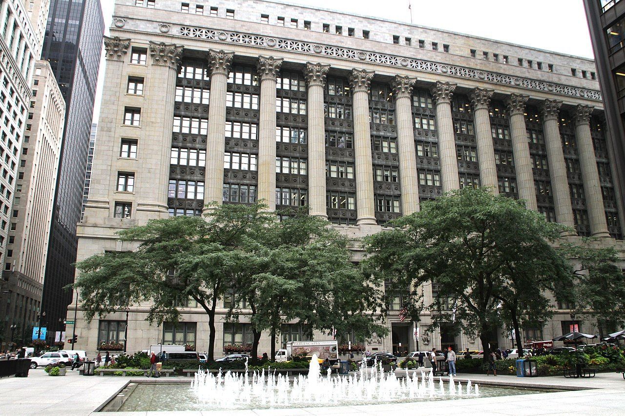 Large, classical building with columns, in a city square, with trees and a fountain in the foreground.