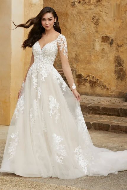 Woman in a white wedding dress with lace detail, a deep V-neckline, and long sheer sleeves, posing outdoors.