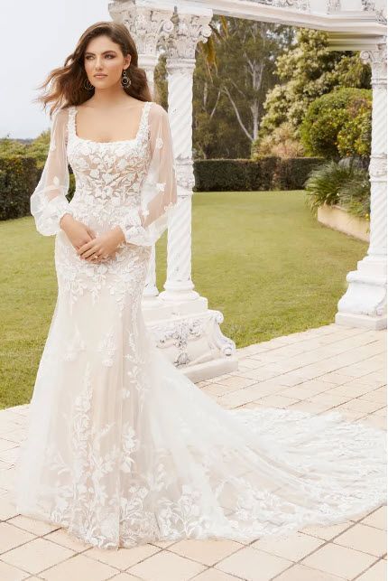 Woman in a wedding dress with sheer sleeves poses outdoors near a white column structure.
