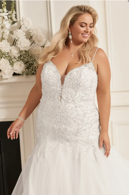 Plus-size bride in a white beaded mermaid gown smiles near a flower arrangement and fireplace.