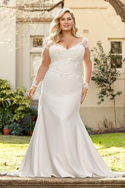 Woman in a white wedding dress with lace sleeves and a mermaid skirt, posing outside a building.