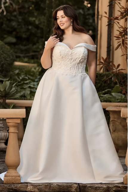 Woman in white off-the-shoulder wedding gown, standing outdoors, smiling, near stone railing.