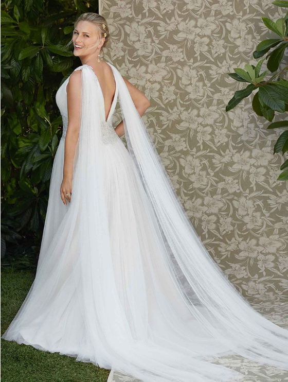 Woman in a white wedding gown with long, flowing cape posing outdoors near greenery and floral backdrop.