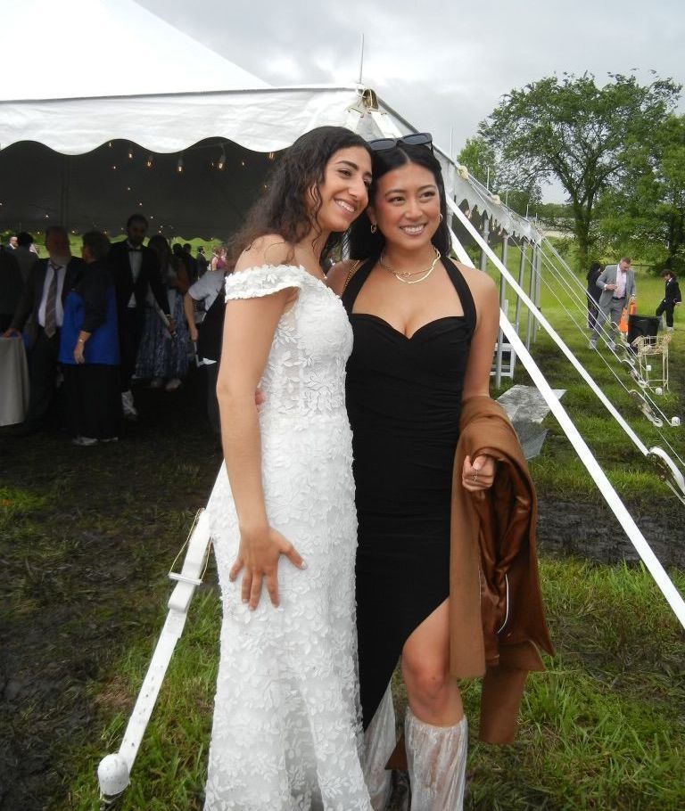 Two women pose for a photo near a white tent. One wears a white dress, the other a black dress and jacket.