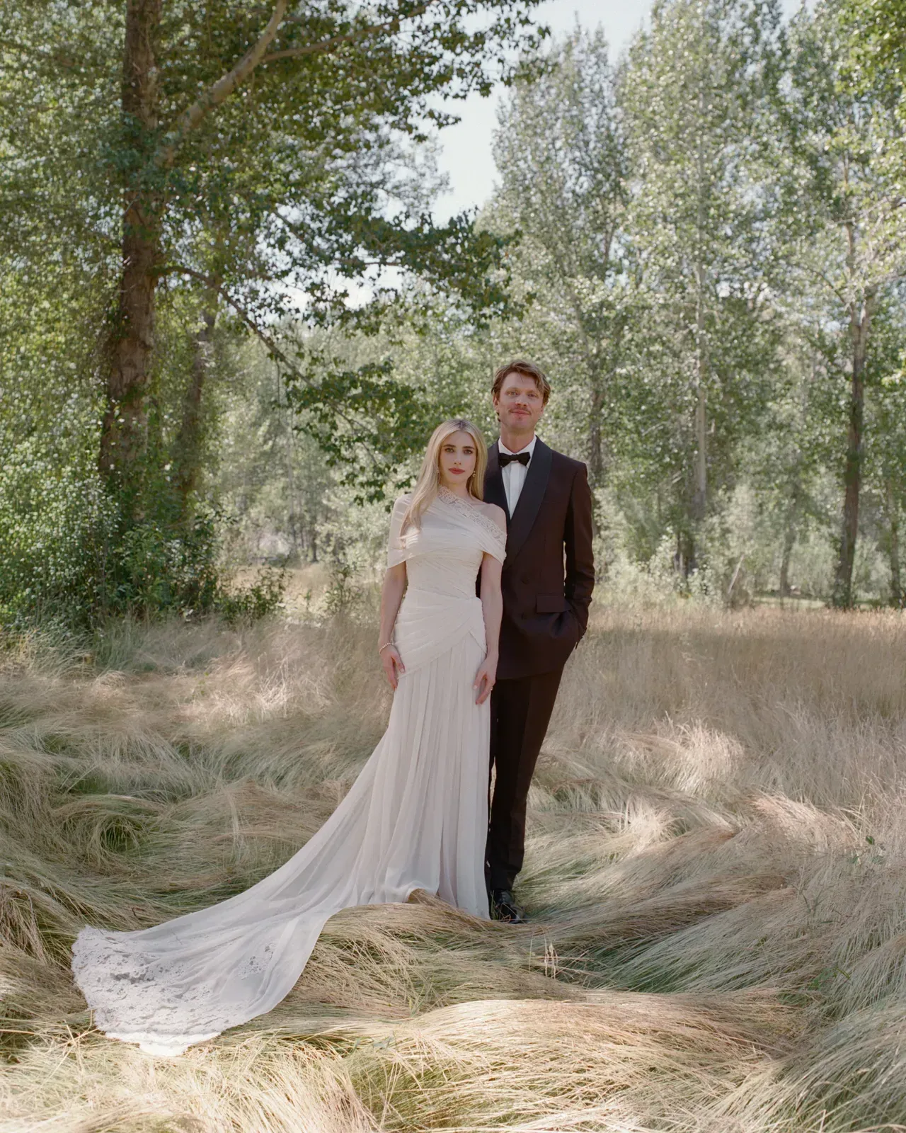 Bride and groom standing in a sunlit woodland, bride in a long white gown with flowing train