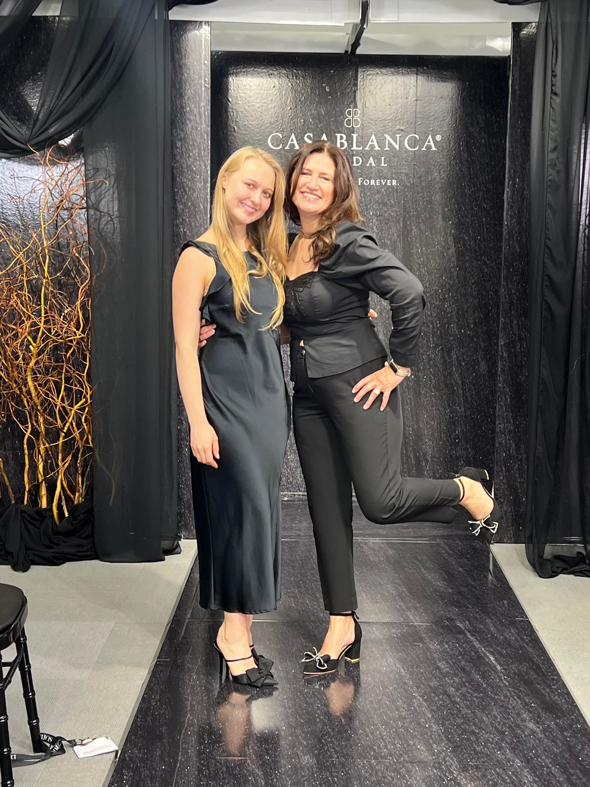 Two women in various outfits posing on a white background. One in a blue jacket and black pants, the other in a black top, skirt, and boots. Fifi and Fivi at Fifi's Bridal