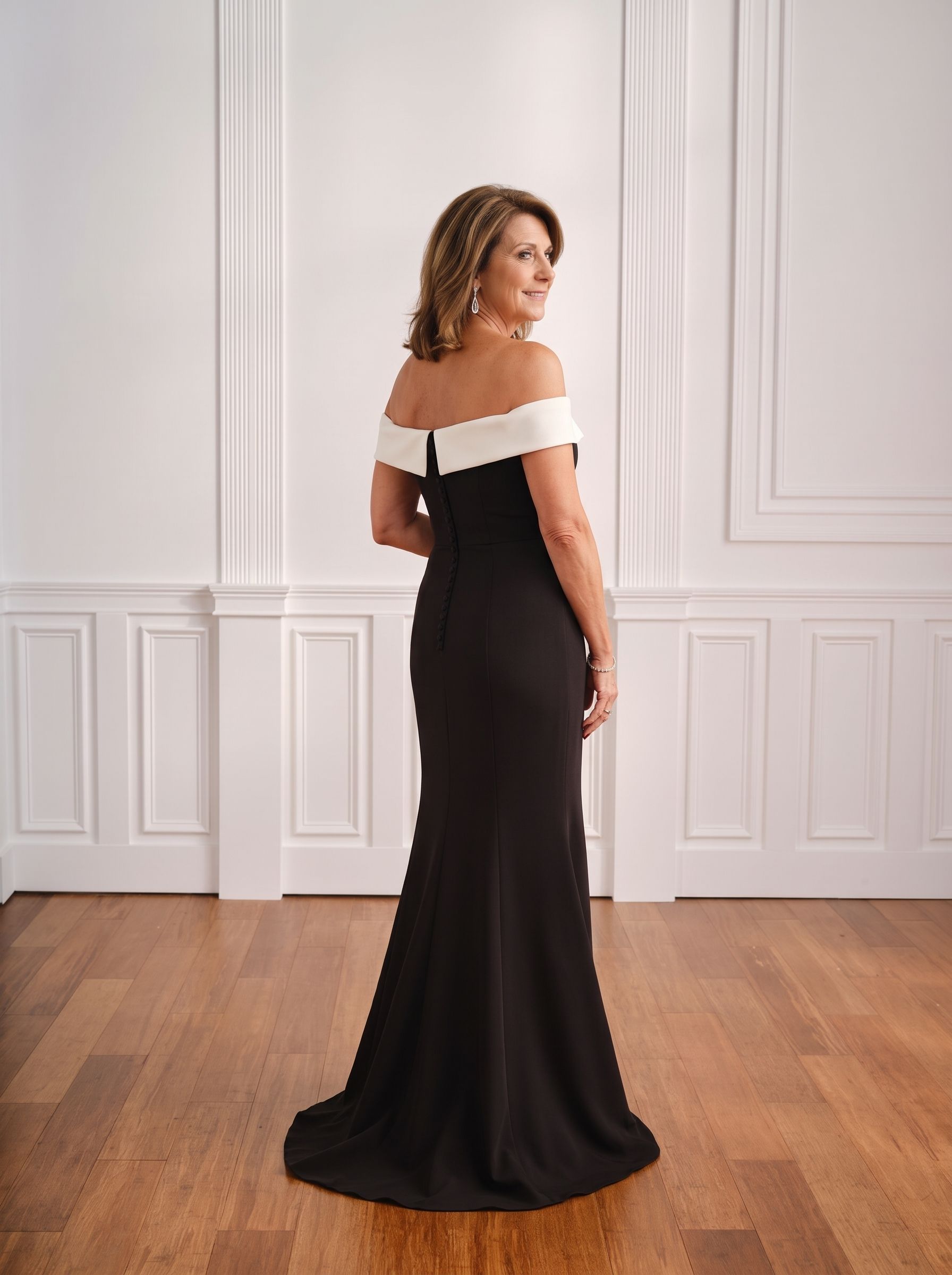 Woman in an off-shoulder black gown standing in a bright room with white paneled walls and wooden floors