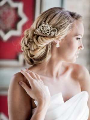 Blonde bride with updo hairstyle and jeweled hairpiece, touching shoulder, against red and white background.