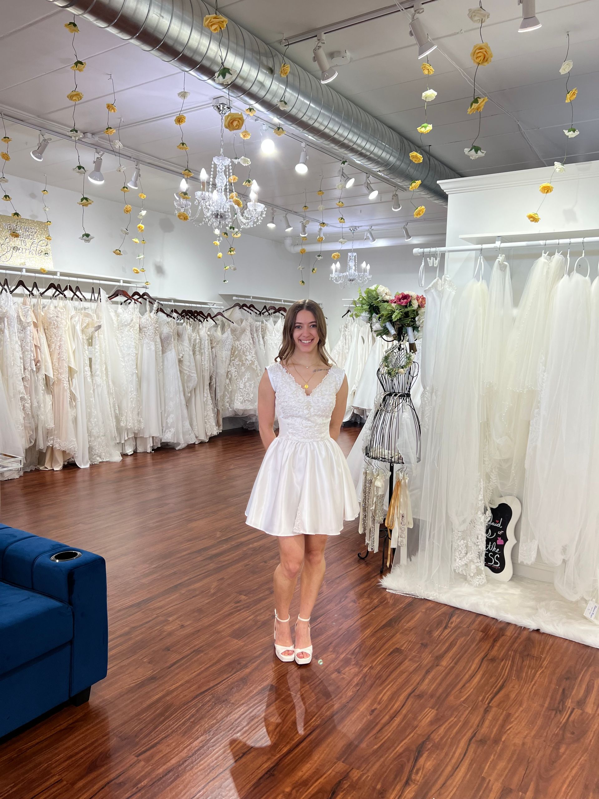 Woman in a white dress standing in a bridal boutique with rows of white gowns and wood flooring