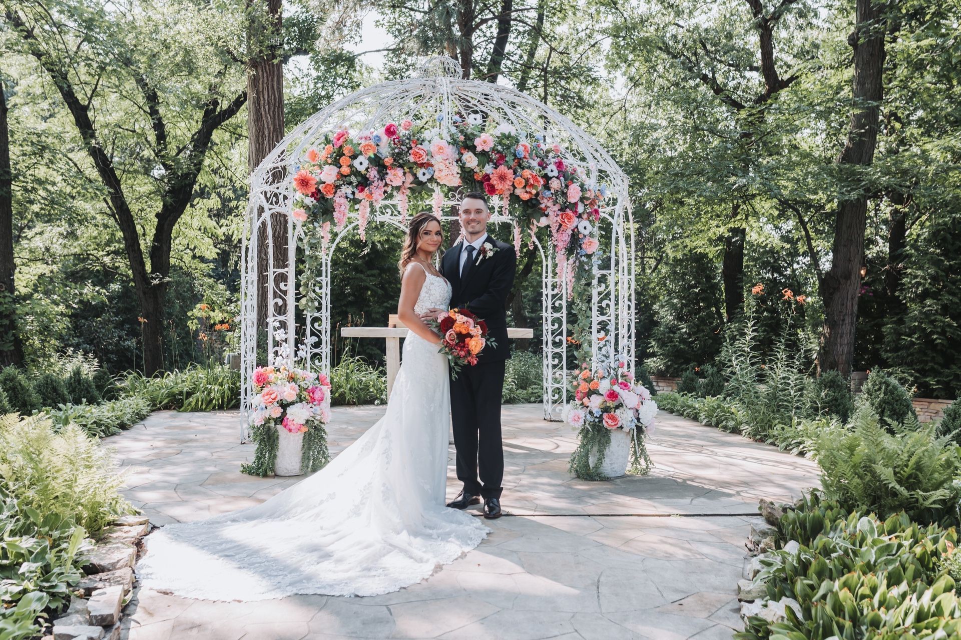 Couple in wedding attire under a floral archway. Outdoor setting with greenery, sunny day.