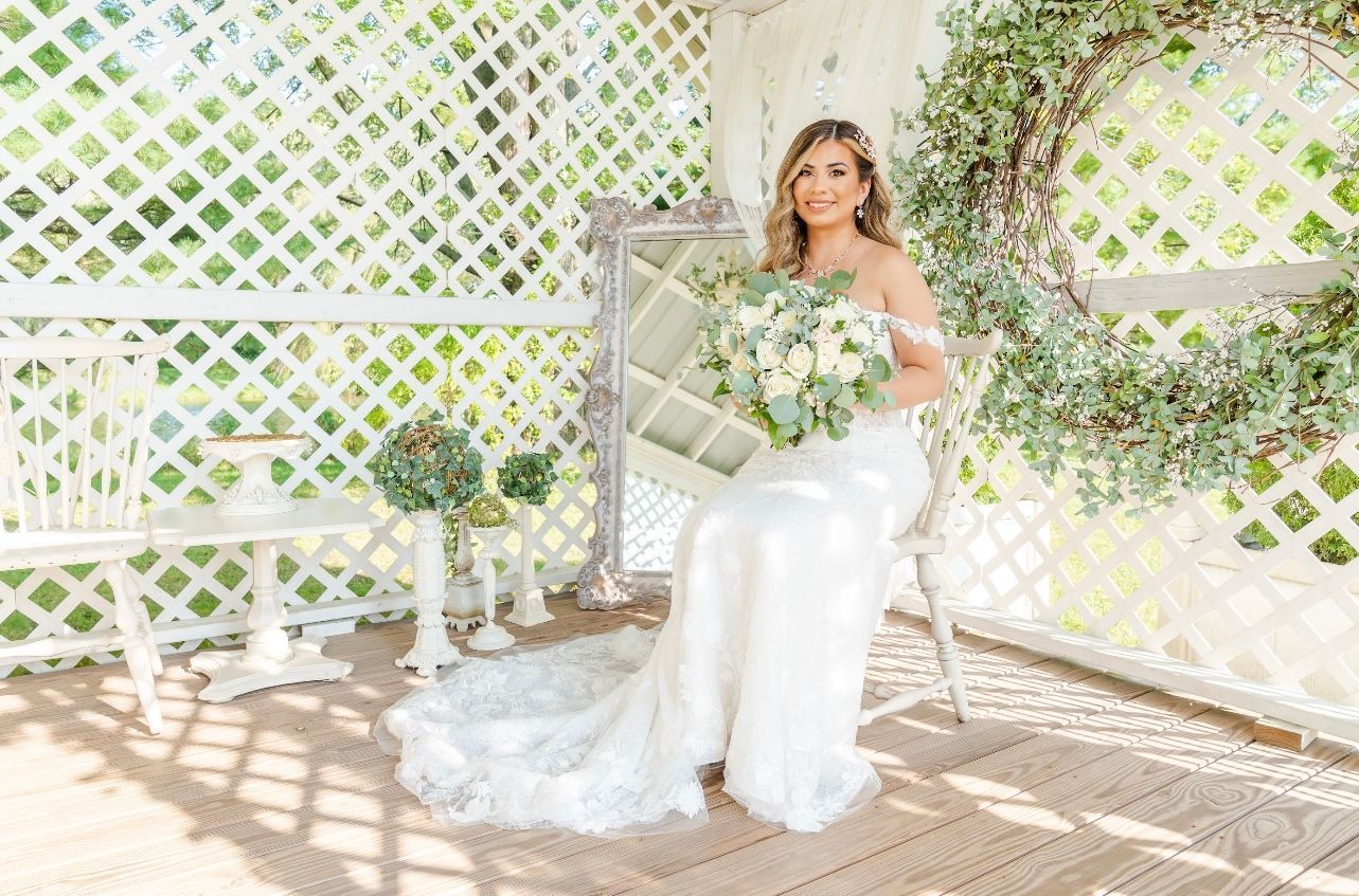Bride in a white dress sits holding a bouquet in a gazebo with a mirror and wreath.