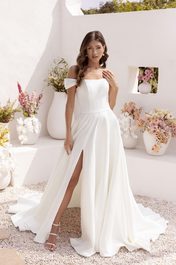Woman in white off-shoulder gown poses outdoors; slit skirt, long train. White walls, flowers, sunny day.