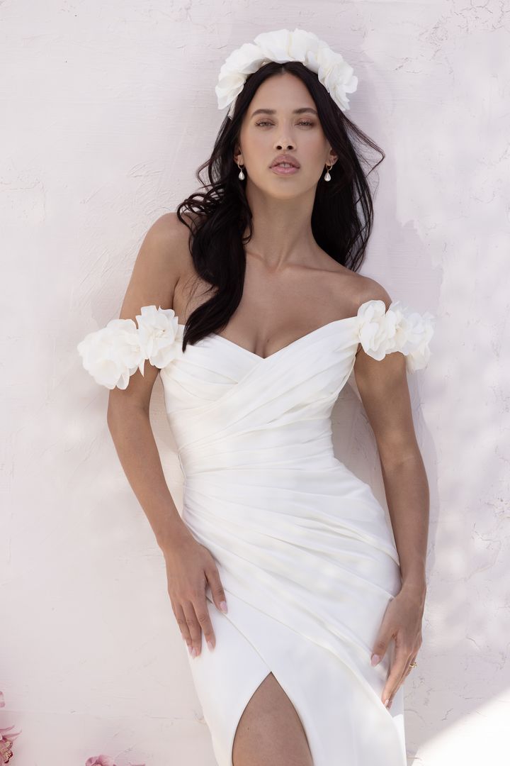 Woman in white off-the-shoulder gown with high slit, standing near flowers and candles against a white background.