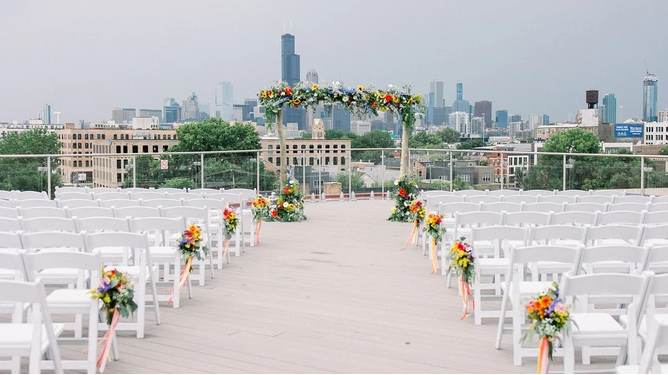Wedding ceremony setup on a rooftop with city skyline. White chairs, floral arch, and colorful floral arrangements.