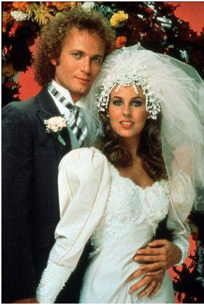 Couple in wedding attire; groom in suit, bride in white gown, veil, embrace, flowers, against colorful backdrop.