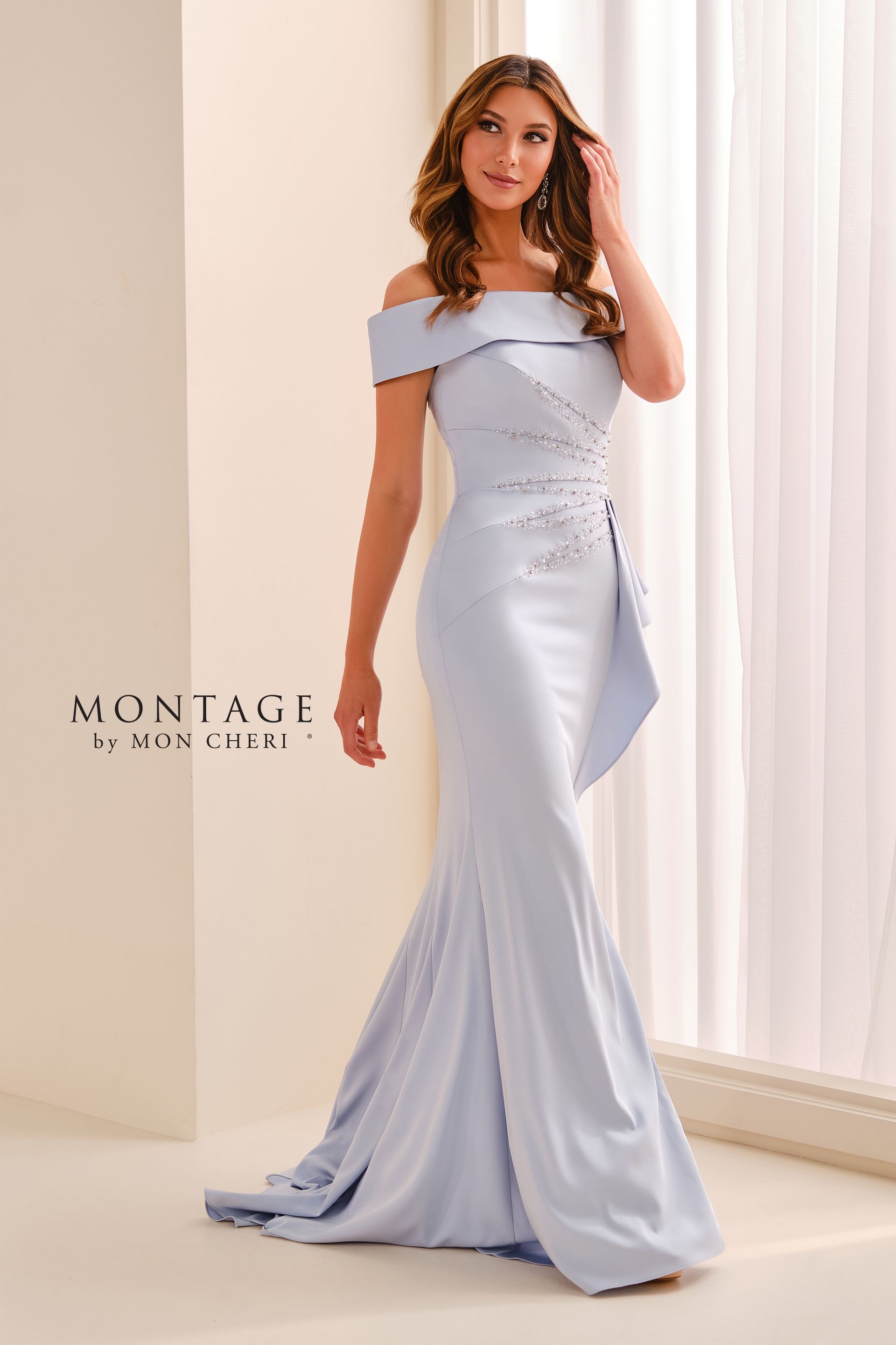 Woman in a light blue off-shoulder dress with beaded details standing indoors by a curtain.