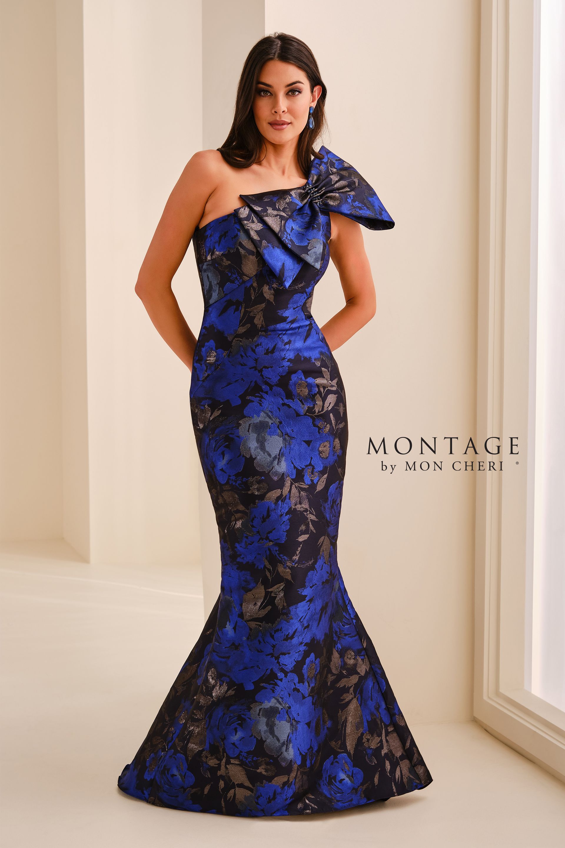 Woman in a blue-and-black floral gown standing in a bright room by a window.