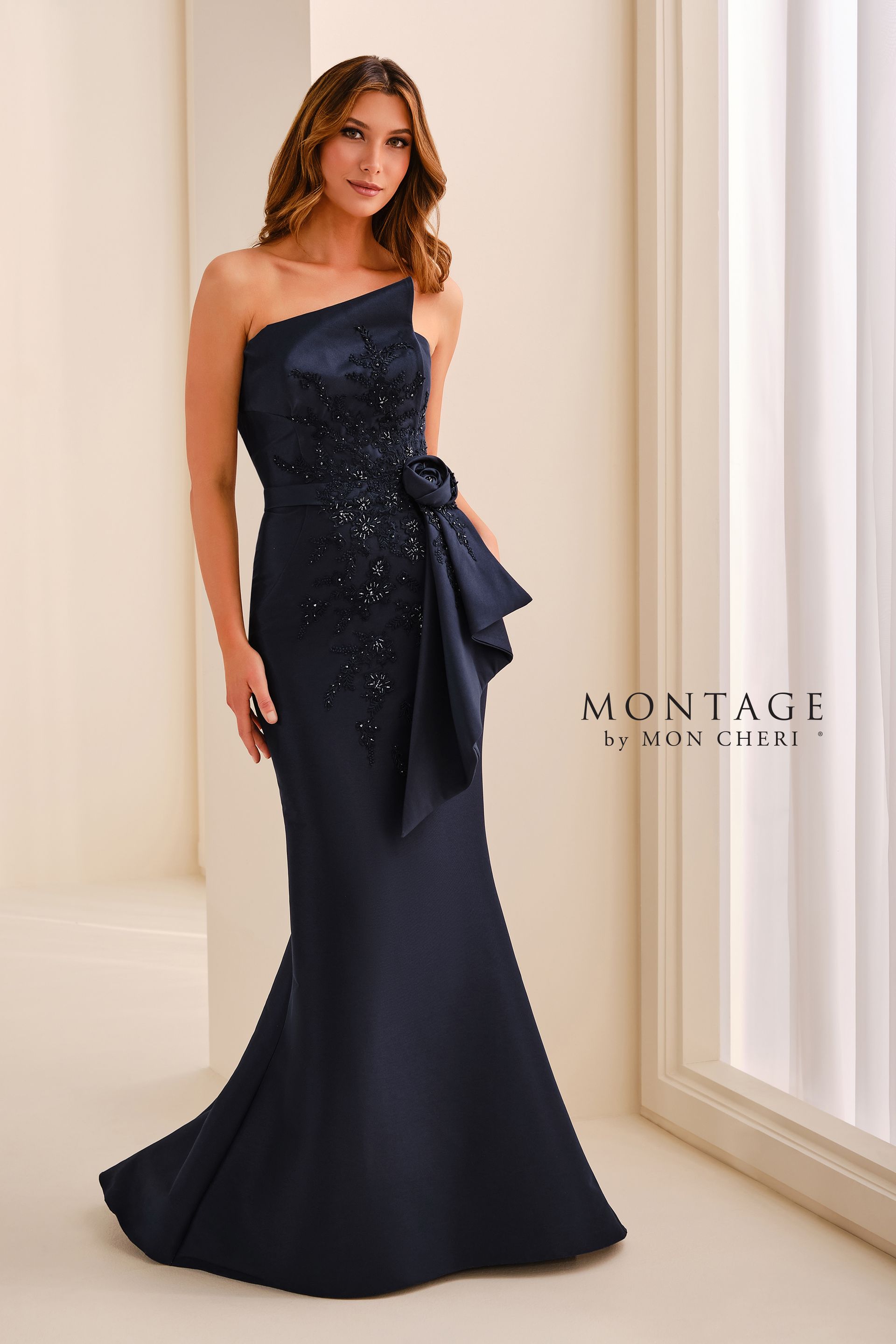 Woman in a navy one-shoulder evening gown with a bow, standing in a bright interior.