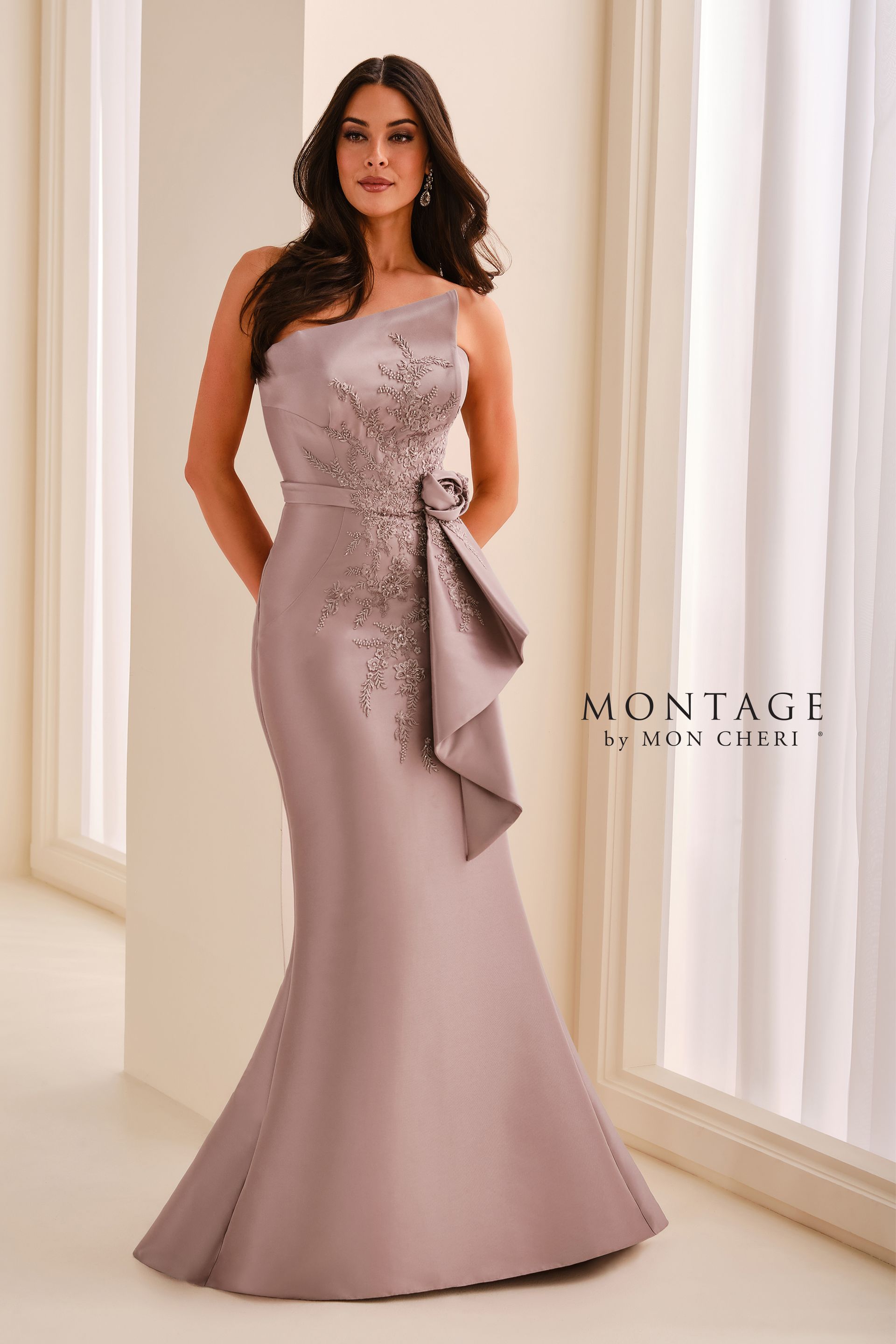 Woman in a navy one-shoulder evening gown with a bow, standing in a bright interior.