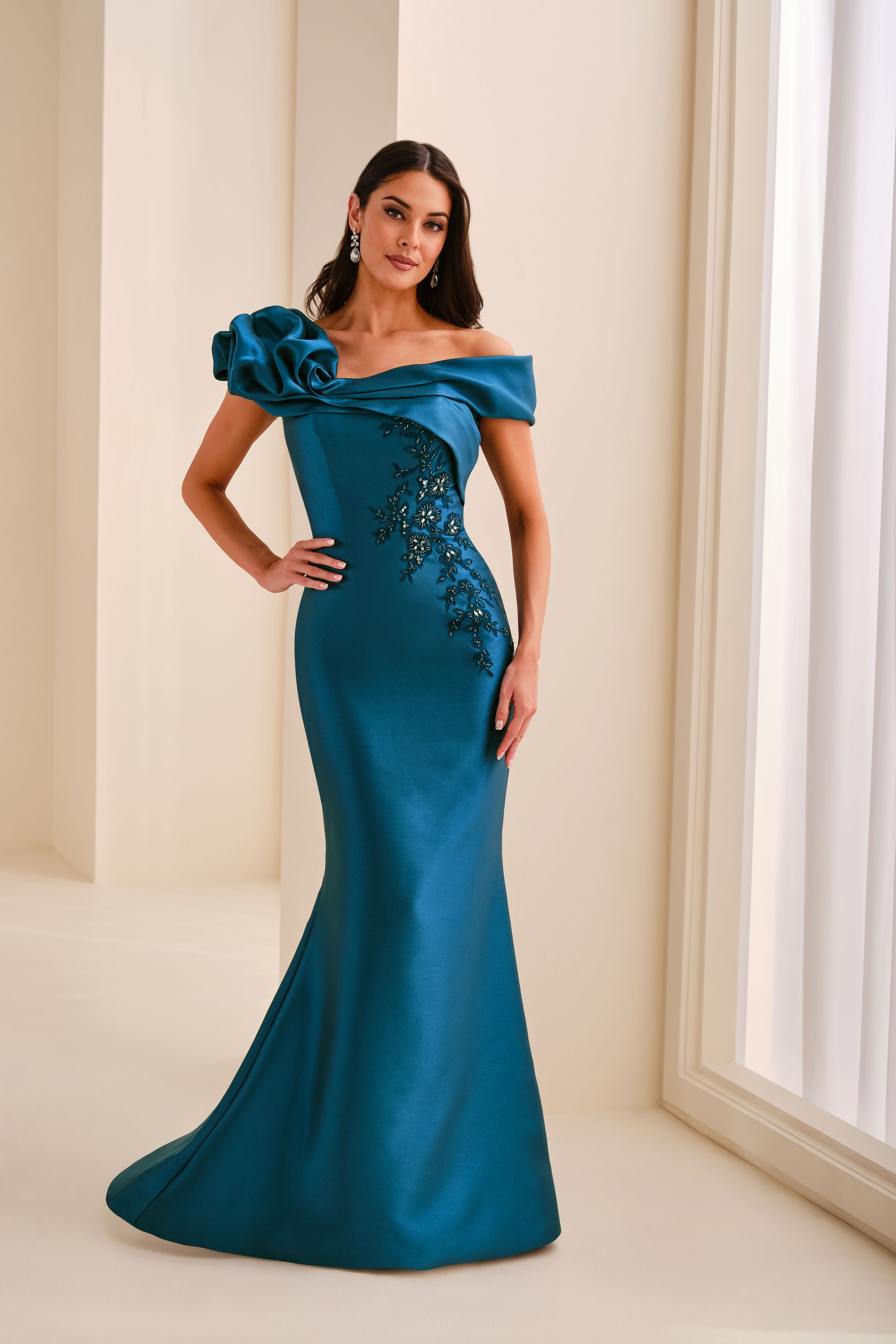Woman in a teal off-shoulder gown standing in a bright room by a window.