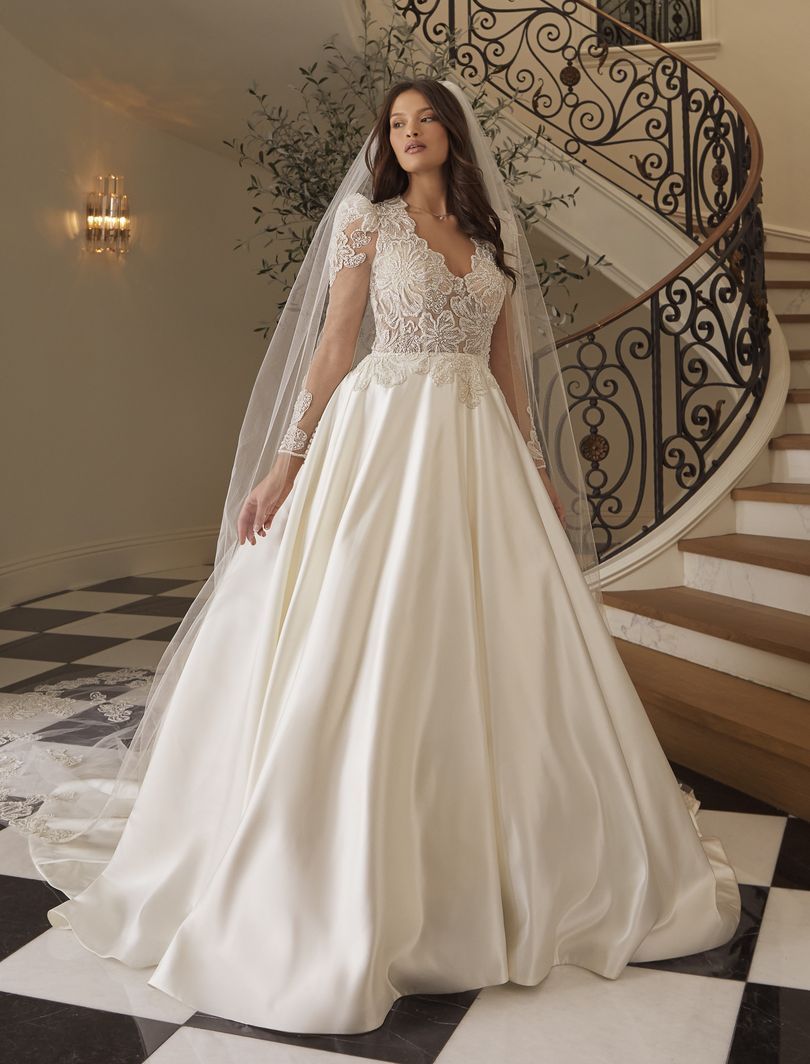 Bride in a white satin wedding dress with lace bodice, wearing a veil, standing by a spiral staircase.