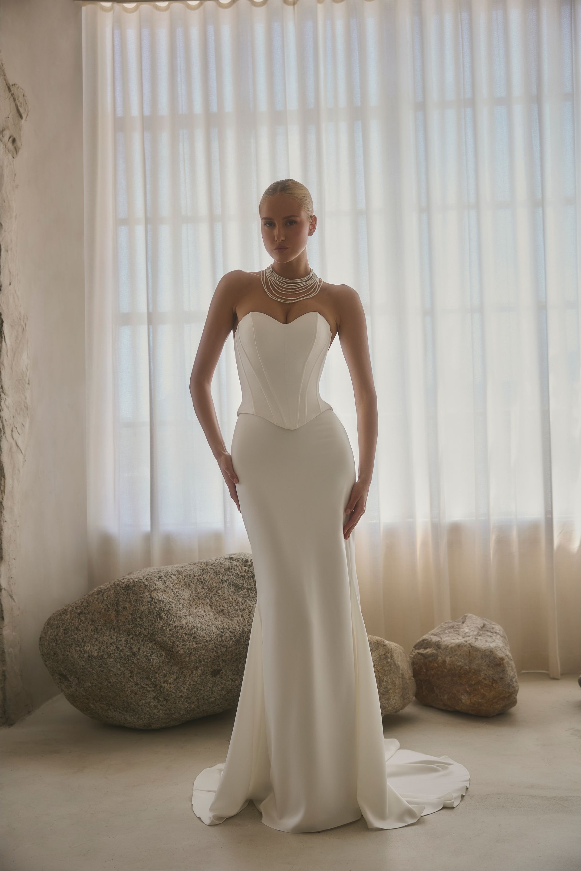 Woman in white strapless wedding gown, standing in front of window, wearing necklace.