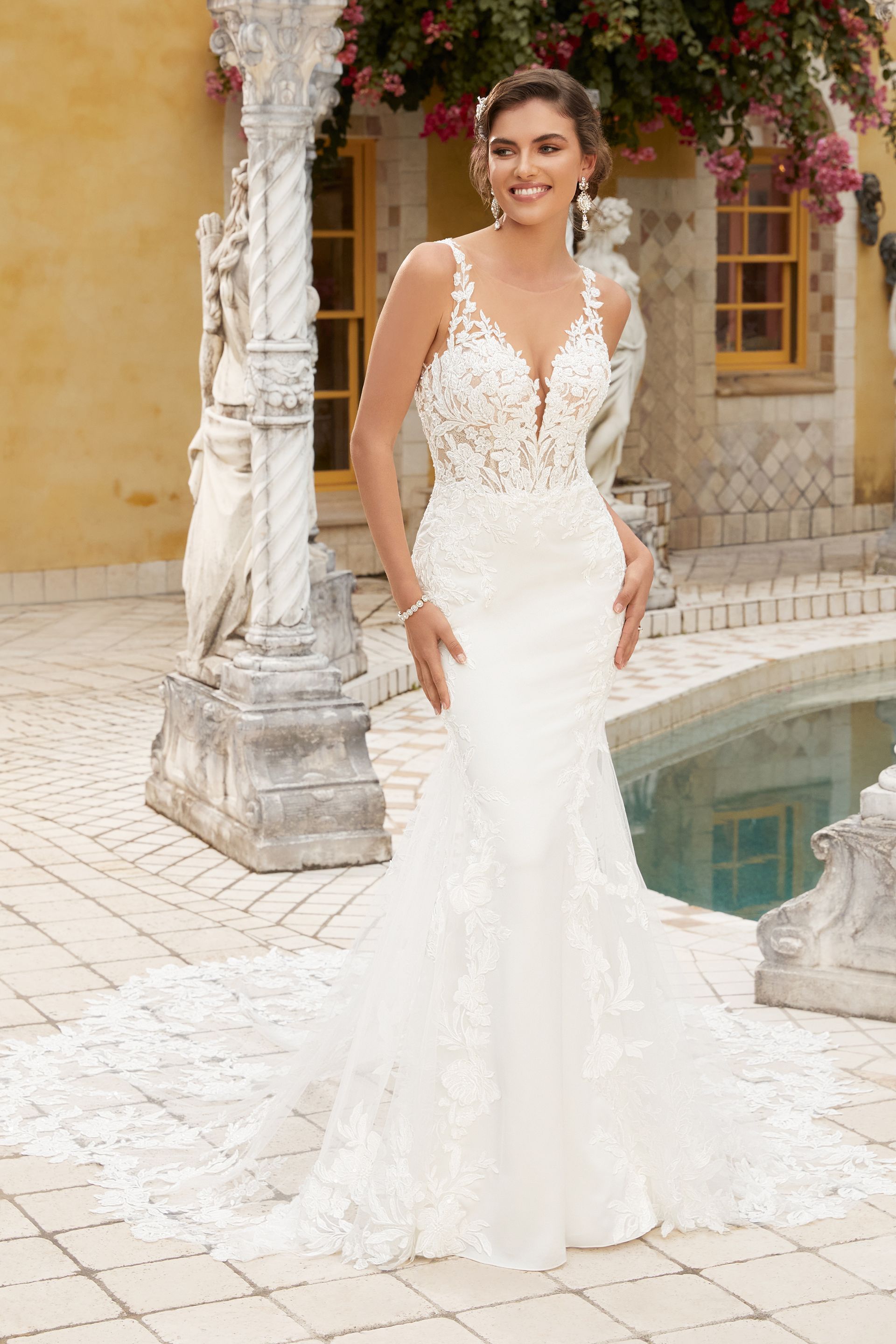 Woman in wedding dress, smiling outdoors near a pool and stone structure. Dress is white, fitted with a train.