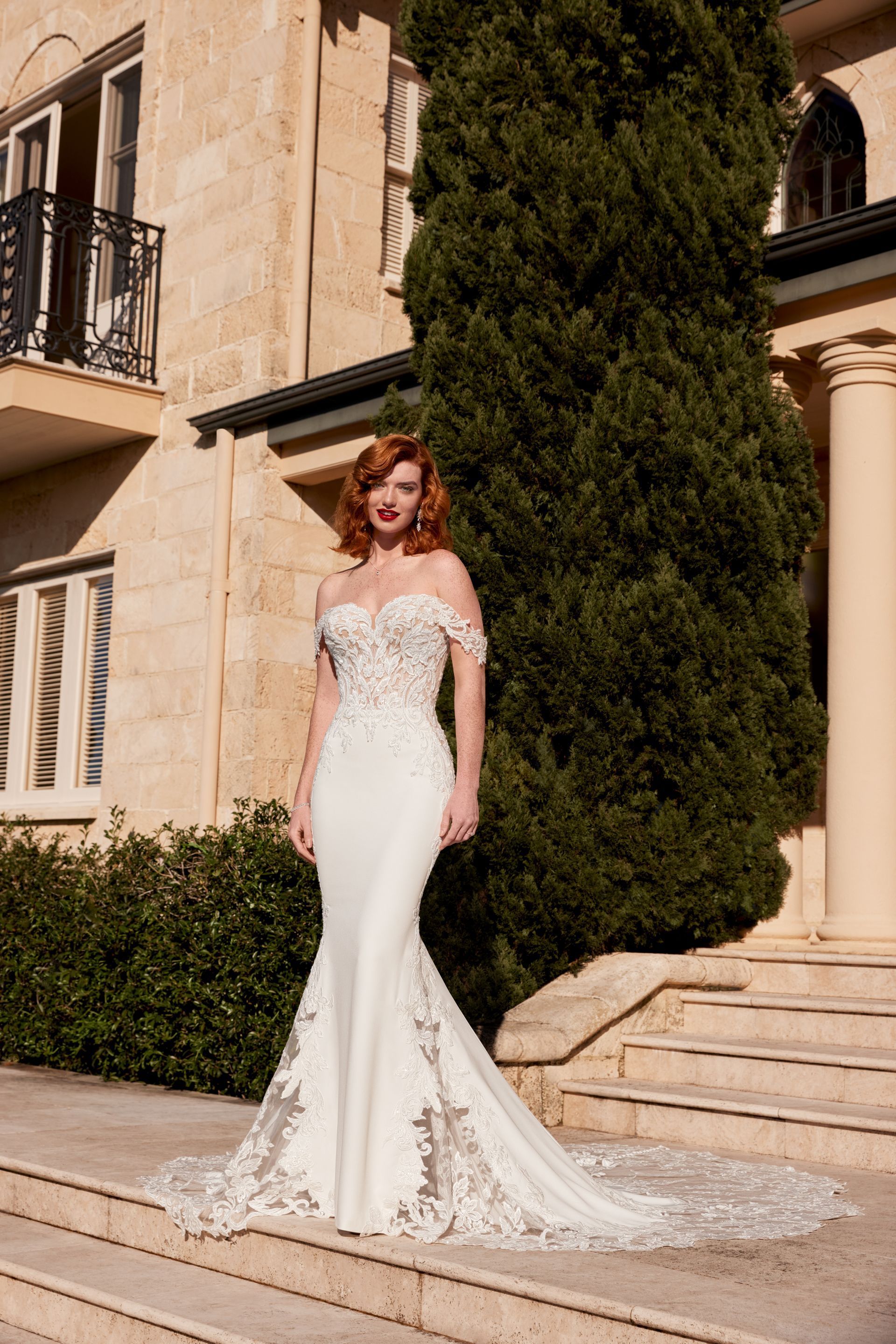 Woman in a white off-the-shoulder wedding dress with a long train, standing outside a stone building.
