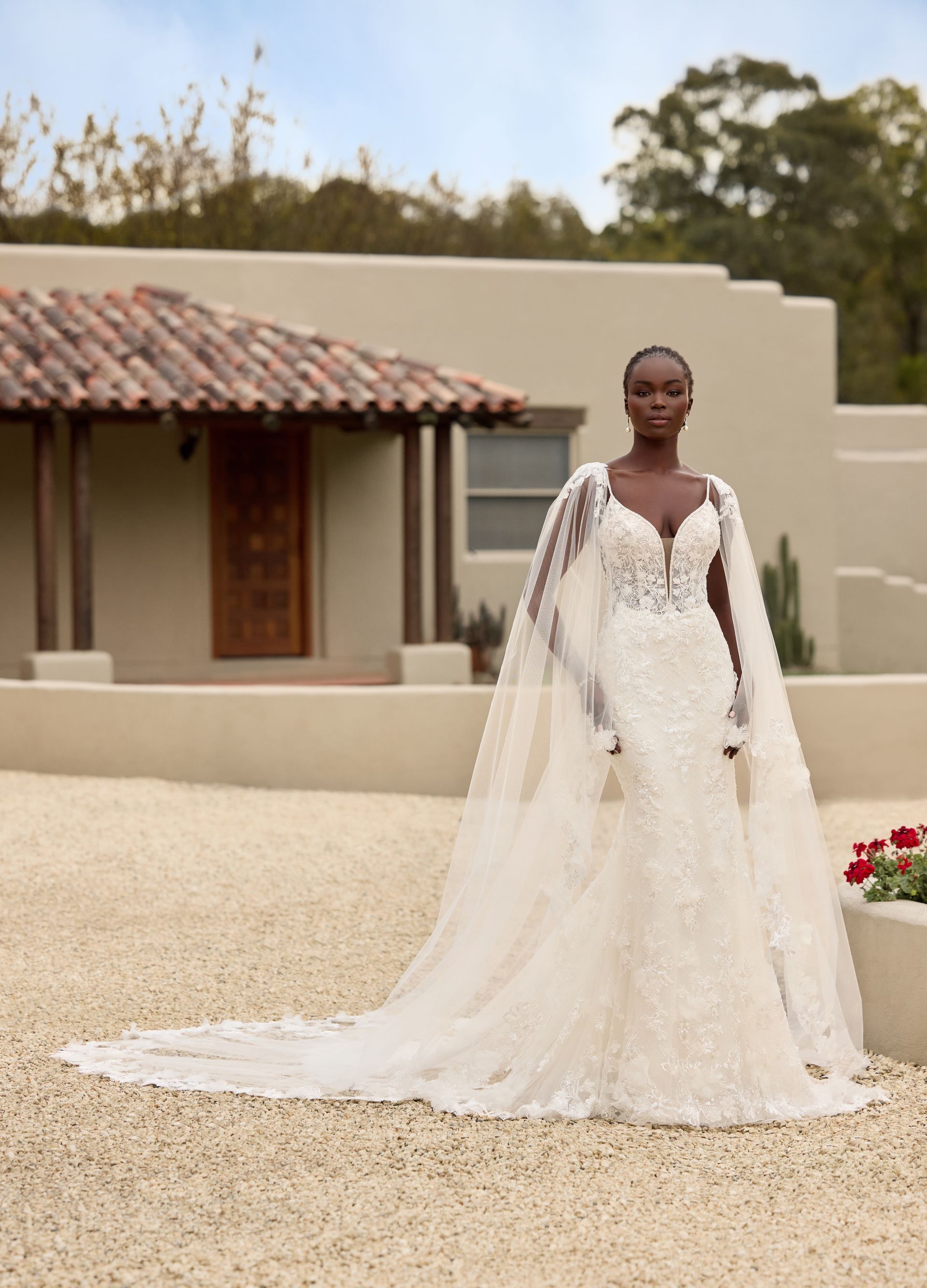 Woman in a beaded wedding dress with cape, standing outdoors in front of a building with a red tiled roof.