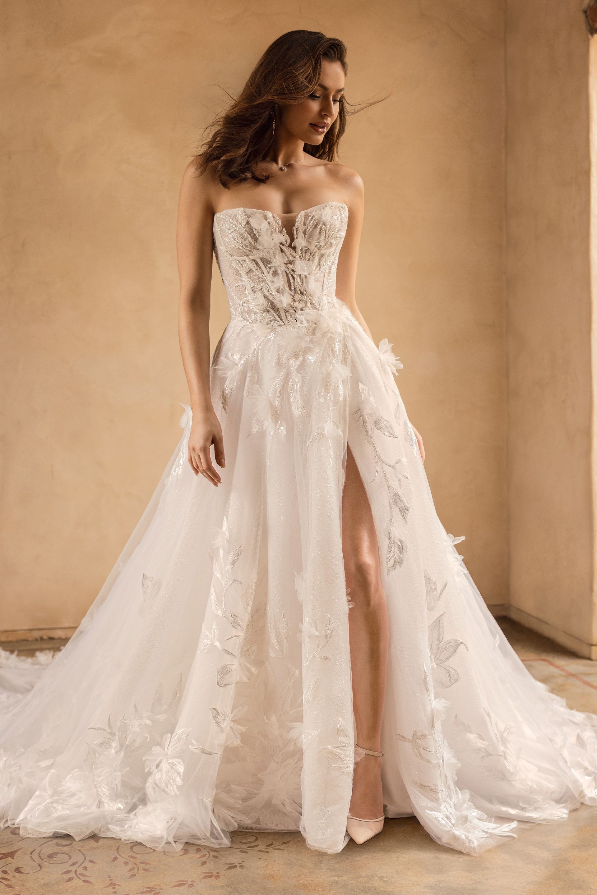Woman in white strapless wedding dress with high slit, floral embellishments, and long train, standing indoors.