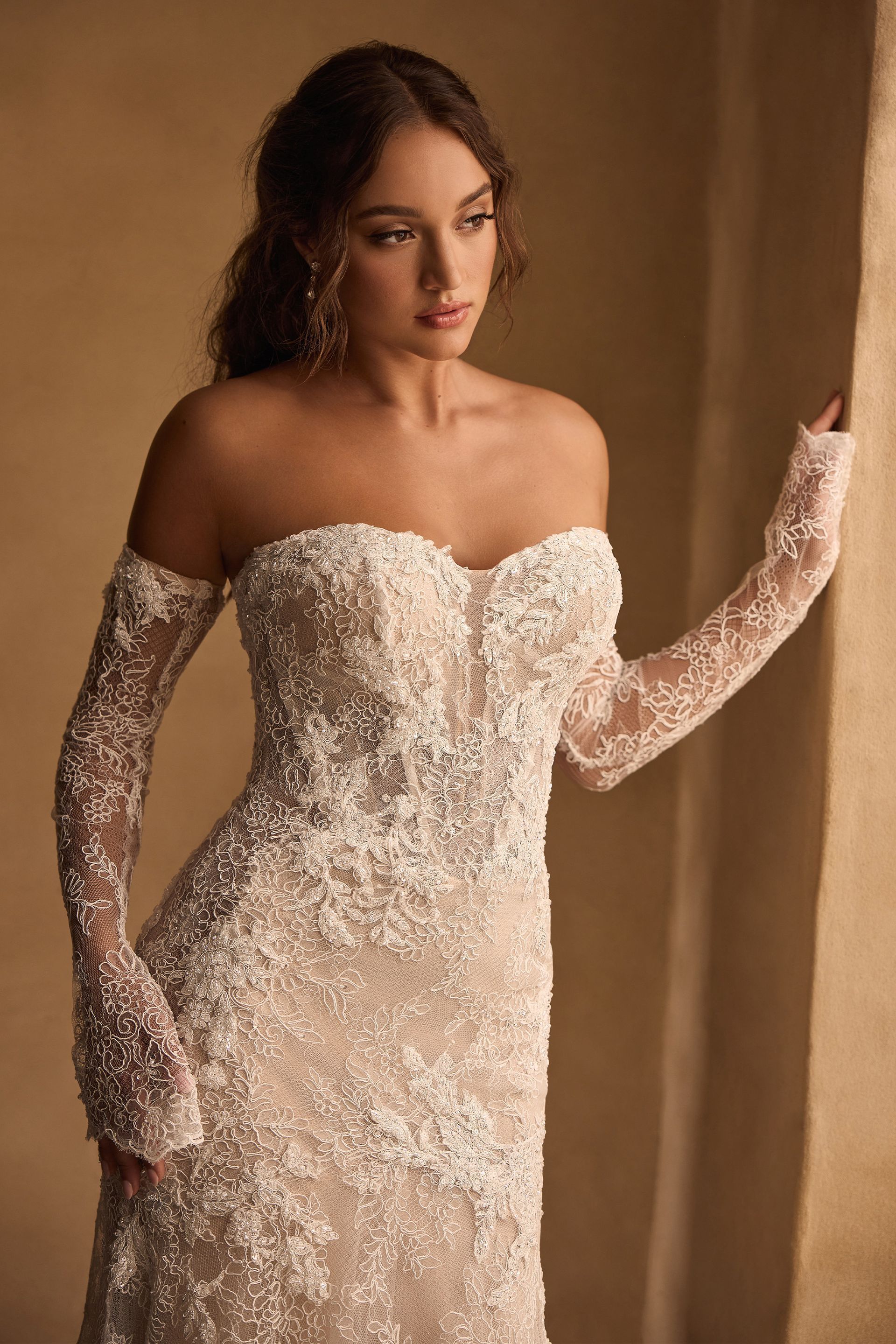 Woman in strapless, beaded wedding dress with lace sleeves, gazing away from window.