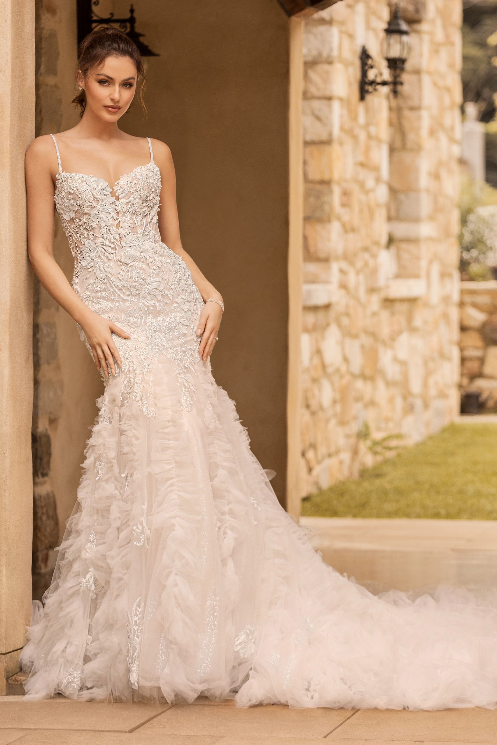 Woman in embellished wedding gown, leaning against stone wall, with a long train.