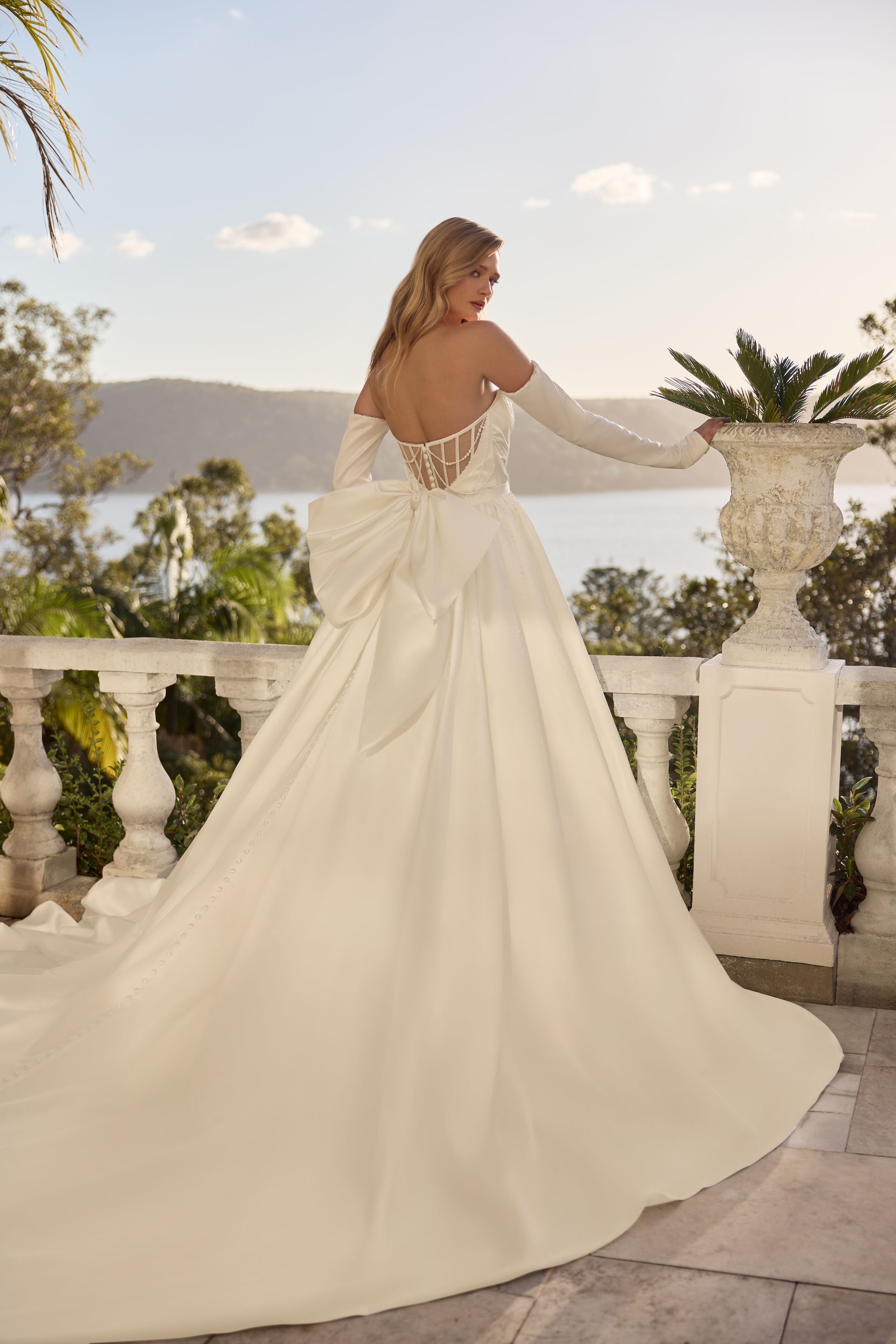 Woman in a white wedding gown with long train, back turned, outdoors.